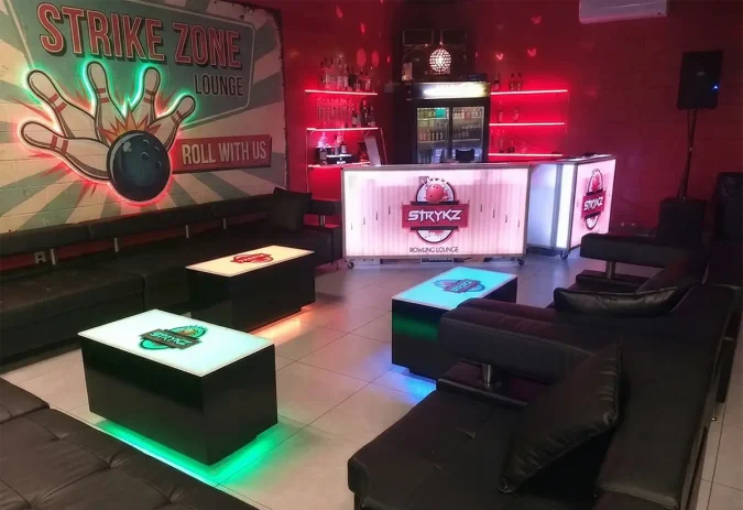 Complete Branded Venue Setup: Custom LED Portable Bars and Cubix Lounge Tables. Bowling alley lounge setup featuring two custom branded portable bars on casters with white LED lighting and bowling alley graphics, surrounded by matching LED lighted cubix cocktail tables.