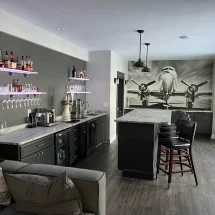 Modern home bar with an aviation mural, featuring two LED floating shelves—one holding liquor bottles and one with hanging wine glass racks, glowing in soft purple.